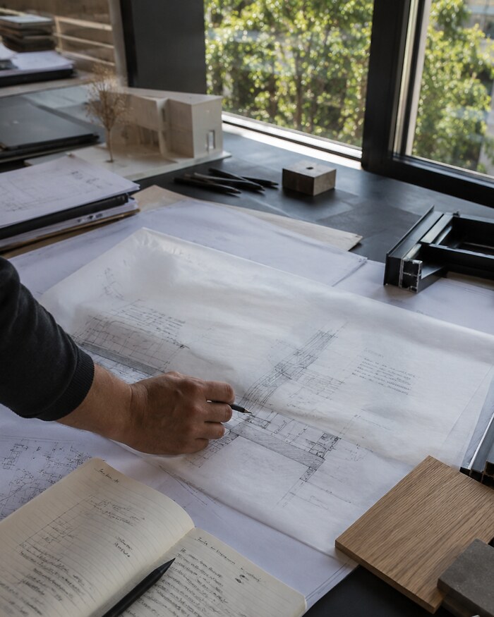 A hand marking up a technical drawing on tracing paper at a drafting table, surrounded by a notebook, scale ruler, material samples and a massing model, with daylight from a studio window
